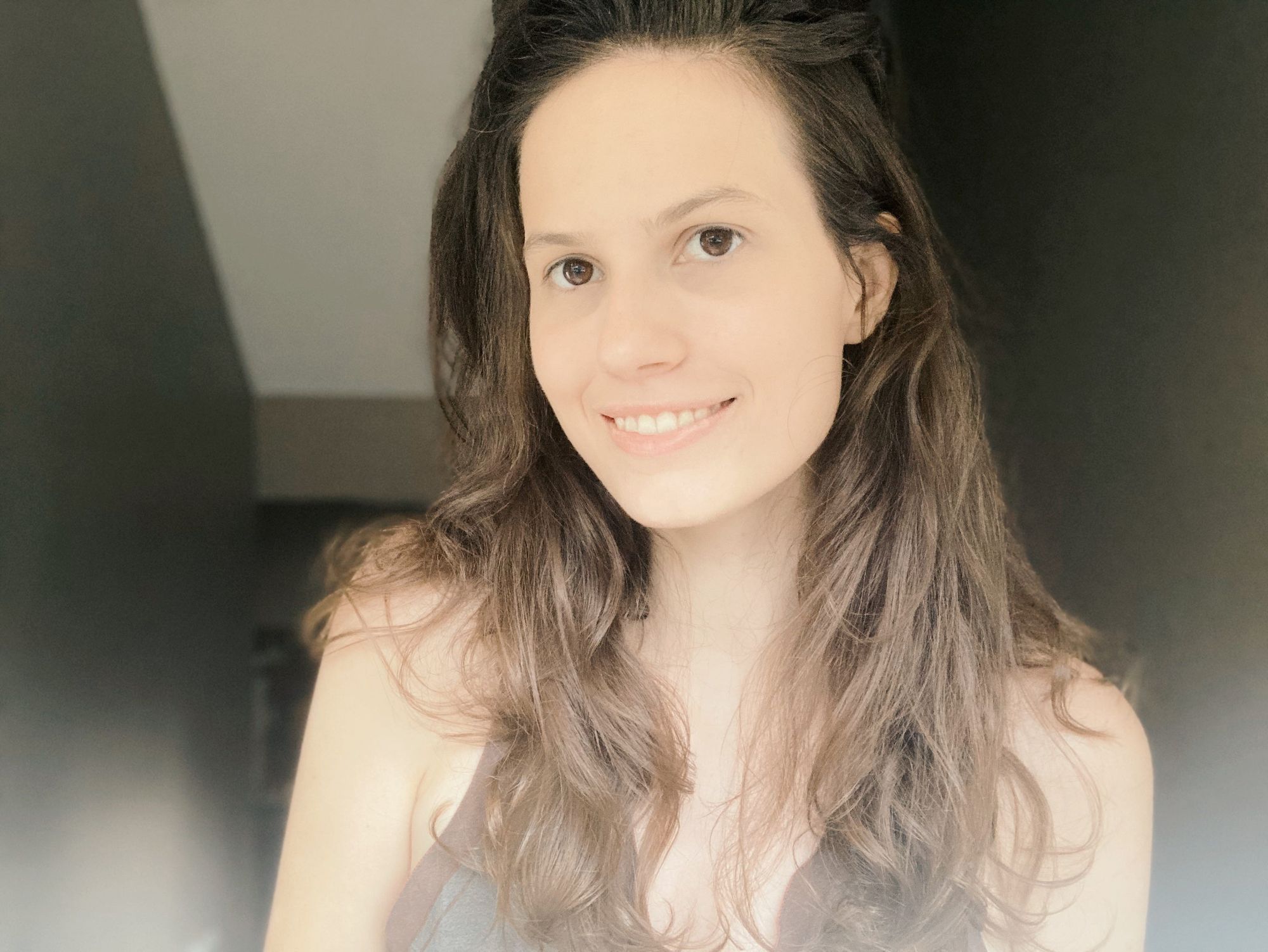 Olivia Muenz, a white woman with long brown hair and brown eyes, is smiling and wearing an olive-colored, sleeveless top. She is in sunlight in front of a darker background.