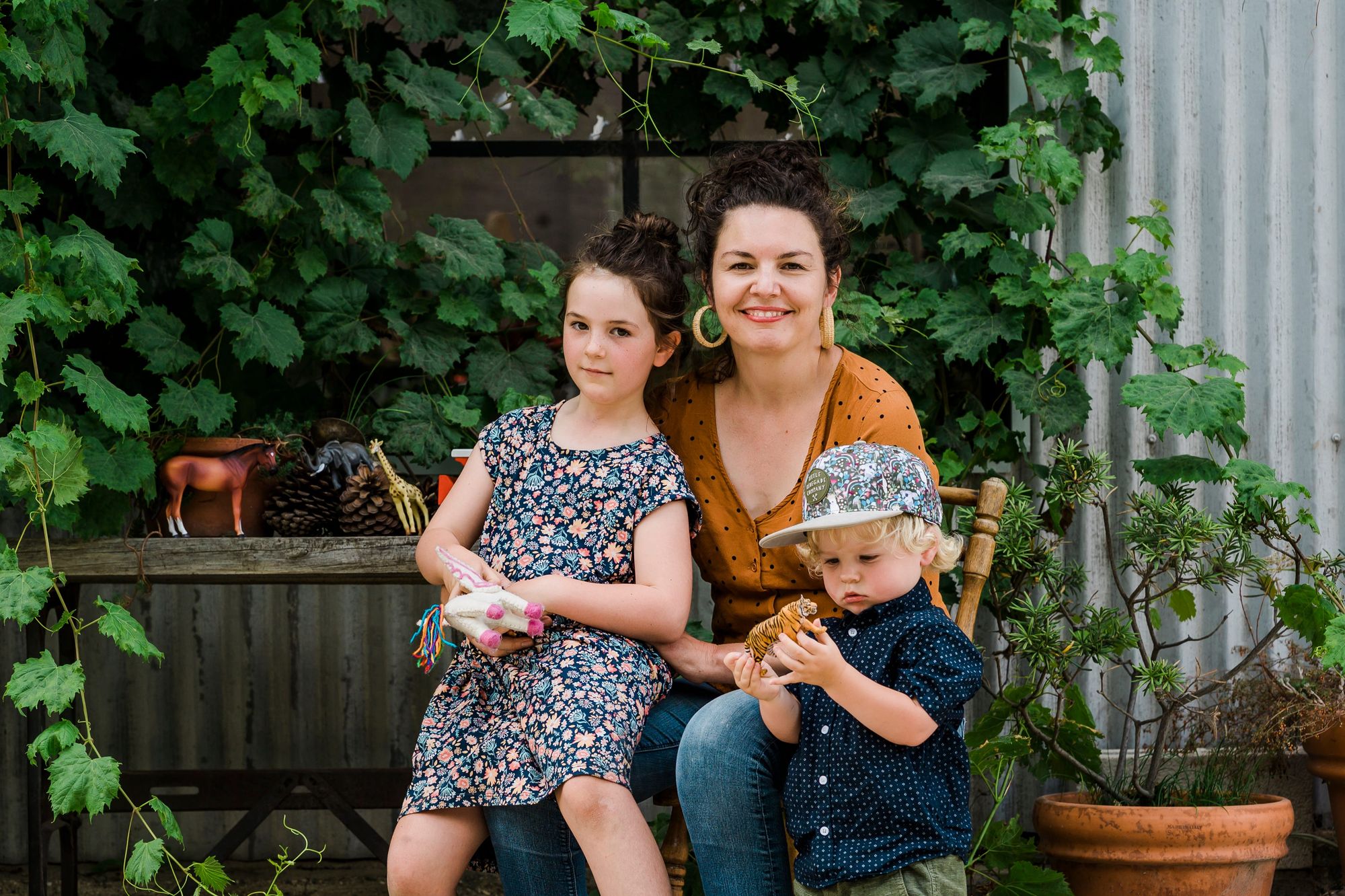The editor, Eliza Hull, sitting in a wooden chair surrounded by her two children both holding toys (a toy horse and a toy tiger). They are in front of a wall filled with greenery. 
