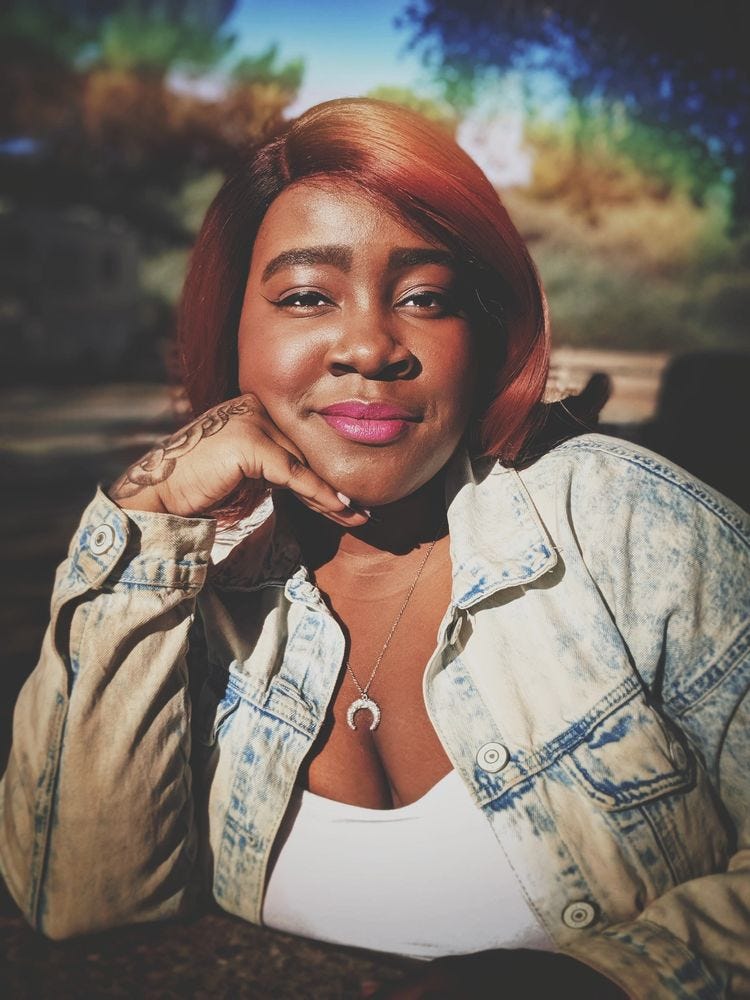 The author, wearing a white shirt and a light-wash denim jacket, sits in a park. Her elbow is resting on the table in front of her and her chin rests on the back of her hand. Photo credit: Cheyenne Ewulu