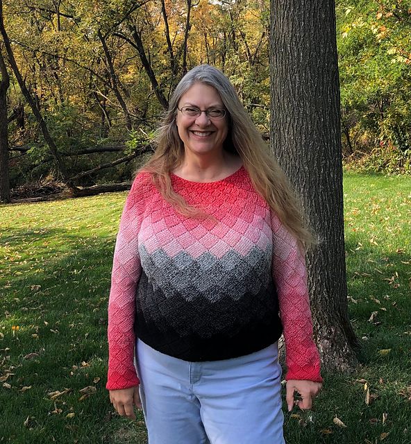 A woman with a bold smile stands showing off her crocheted pink, grey, and black gradient sweater.