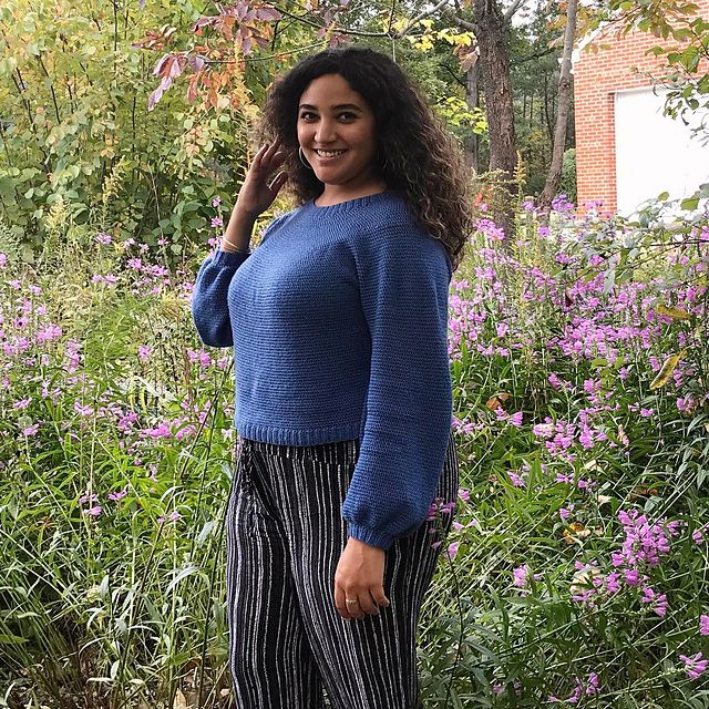 A woman smiles confidently at the camera, standing in a field of flowers and wearing her newly-finished crocheted sweater.