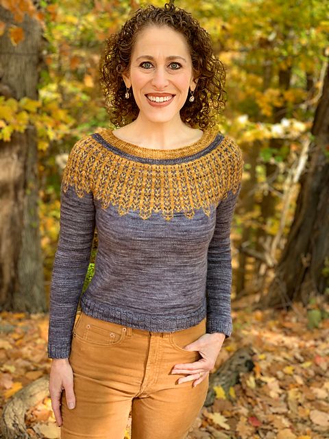 A smiling woman in a grey sweater with a gold colorwork yoke motif that pays homage to Ruth Bader Ginsberg's collars.