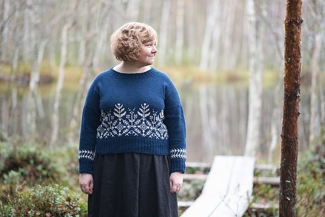 A woman stands in a forest, near a lake, wearing a rich blue and white colorwork sweater.