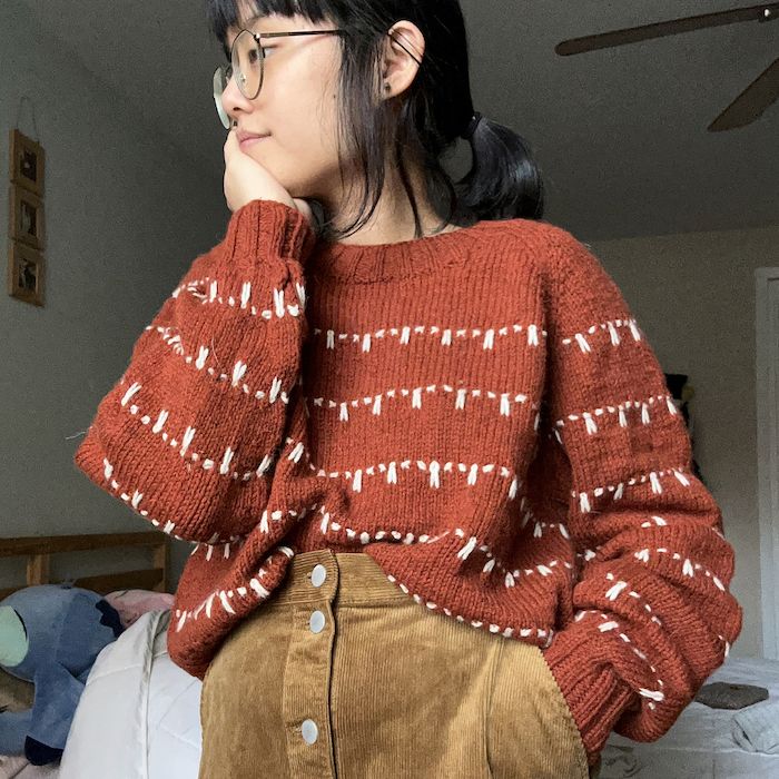 A woman in a cozy, oversized rust-colored knit sweater with a cream striped detail, her dark hair in a ponytail, stands with her hand on her chin, looking to the left of the frame.