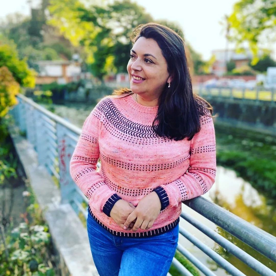 a woman stands near a stream of water, looking to the side of the camera and smiling. her pullover sweater is bright pink with deep plum accent stitches.