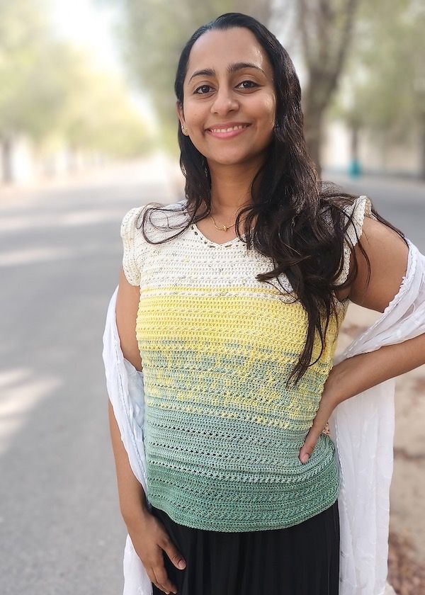 A smiling woman looks at the camera with her hand on her hip; she is wearing a crocheted tank with ruffles at the armholes, worked in a gradient of cream, yellow, and sage greens.