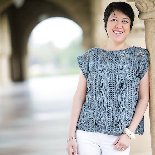 A woman leans against a column, smiling broadly, wearing a crocheted tank with vertical eyelet details.