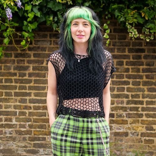 A Raveler stands in front of a brick wall, hands in the pockets of her green plaid pants, wearing a knit mesh tank worked in black.