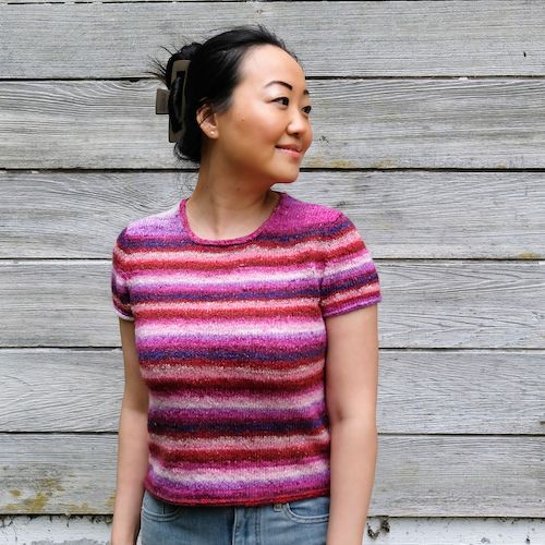 A woman stands in front of a wooden wall, looking to the right of camera with a smile. She's wearing a brightly striped pink sweater in shades of pink.