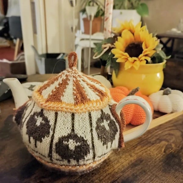 A white ceramic teapot with a cozy made to look like a carousel with sheep "seats" is on a table that also has yarn pumpkins and a yellow vase with sunflowers.