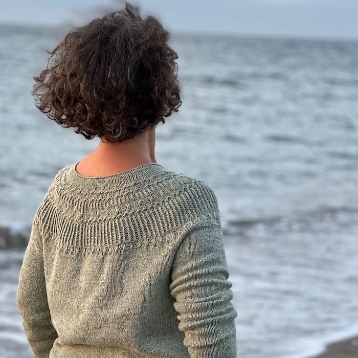 Standing facing the sea, a woman wears a textured grey sweater with a circular yoke.