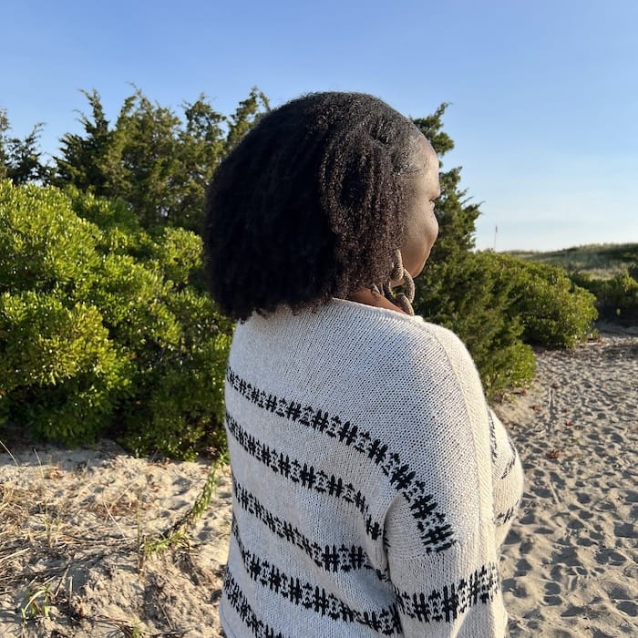 A woman stands in a sandy spot by the trees in a soft cream sweater with bold dark colorwork stripes.