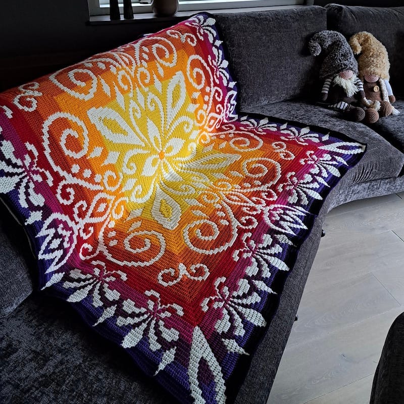 a grey couch is draped with a bold crocheted blanket, radiating in sunset colors from a yellow center, with a white stranded design contrasting
