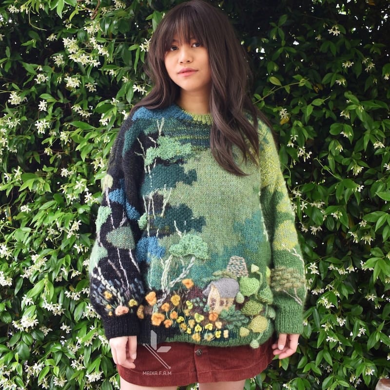 A knitter stands against a green flowery wall; her sweater has a richly textured scene worked into it, with trees, a cottage, and flowers.