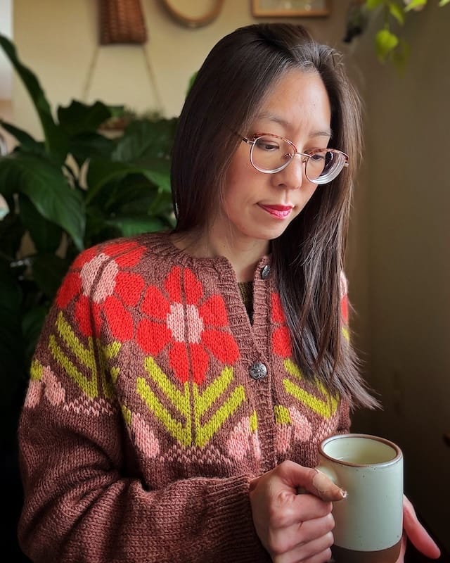 A crafter looks peacefully at a mug of tea, wearing a cozy cardigan with large bold red flowers knit into the yoke.