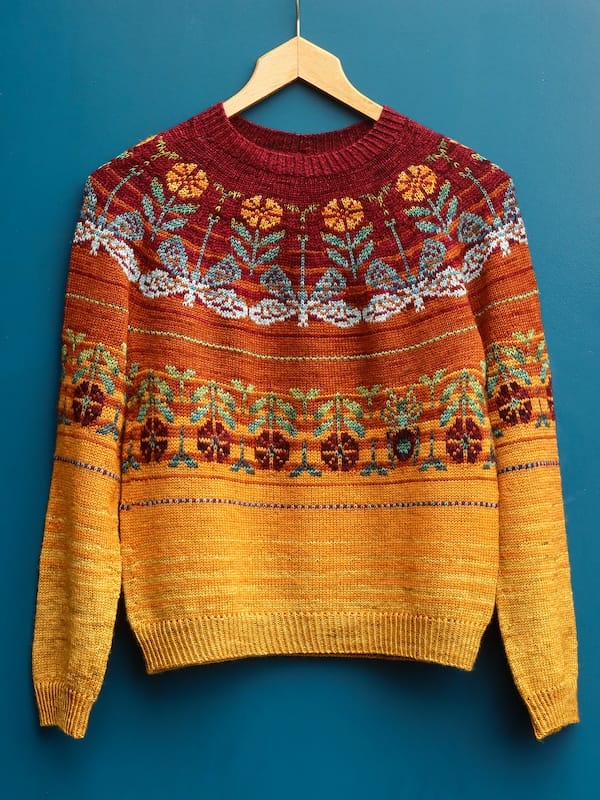 A sweater in bold autumnal colors, with stranded colorwork insects and flowers, hangs from a hanger against a rich teal background.