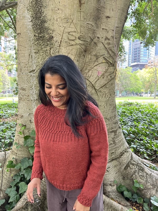 A woman stands in front of a tree, smiling, looking down. Her handknit sweater is a rich deep orange, with a circular textured yoke.