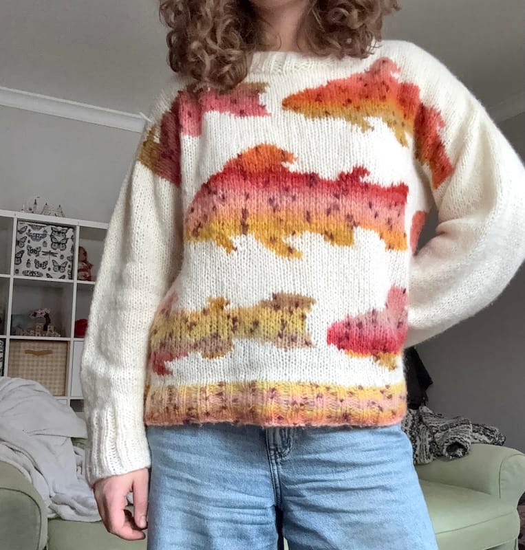 A crafter stands with one hand on hip, showing off their handmade sweater in ivory with giant trout designs worked throughout.