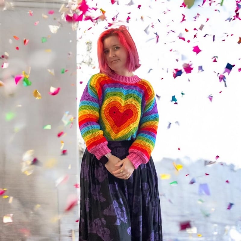A crafter poses among colorful confetti, wearing a rainbow colored crocheted sweater with a heart motif on the front.