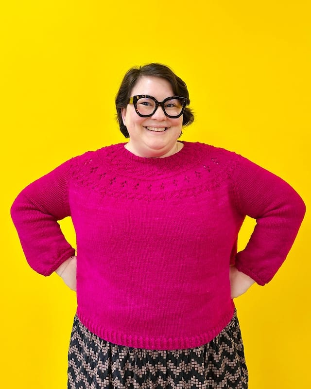 A woman stands, hands on hips, in a bright fuschia sweater against a vibrant yellow wall.