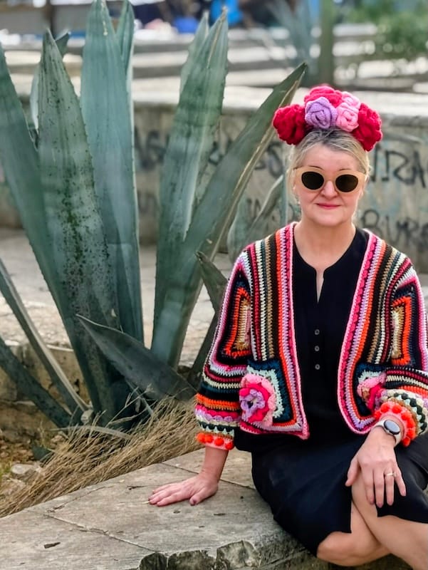 A crocheter sits on a low wall in front of a large desert plant; she wears a brightly striped crocheted cardigan with rosettes on the pockets.