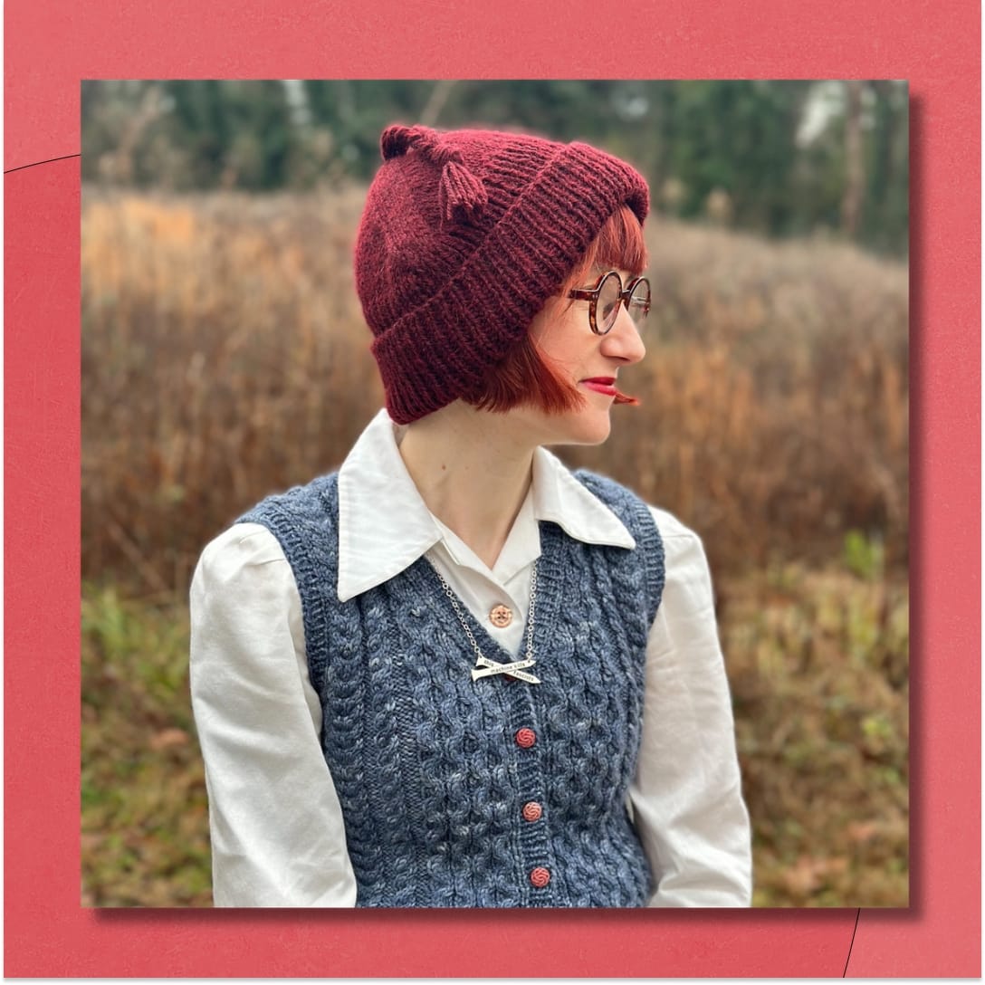 A woman wearing a burgundy handknit hat with a tassel looks to the right. She is standing outdoors in a field with brown and green grasses.