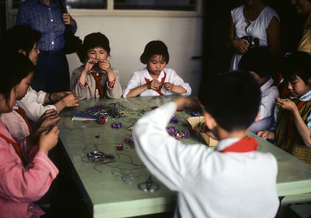 1979 color photograph of several Chinese children intently making beaded crafts around a light green table. On the table are colorful string and beads, scissors, and other supplies. The kids wear a range of outfits, but all have matching red bandana ties. A few adults stand against the wall. Only their torsos are pictured.