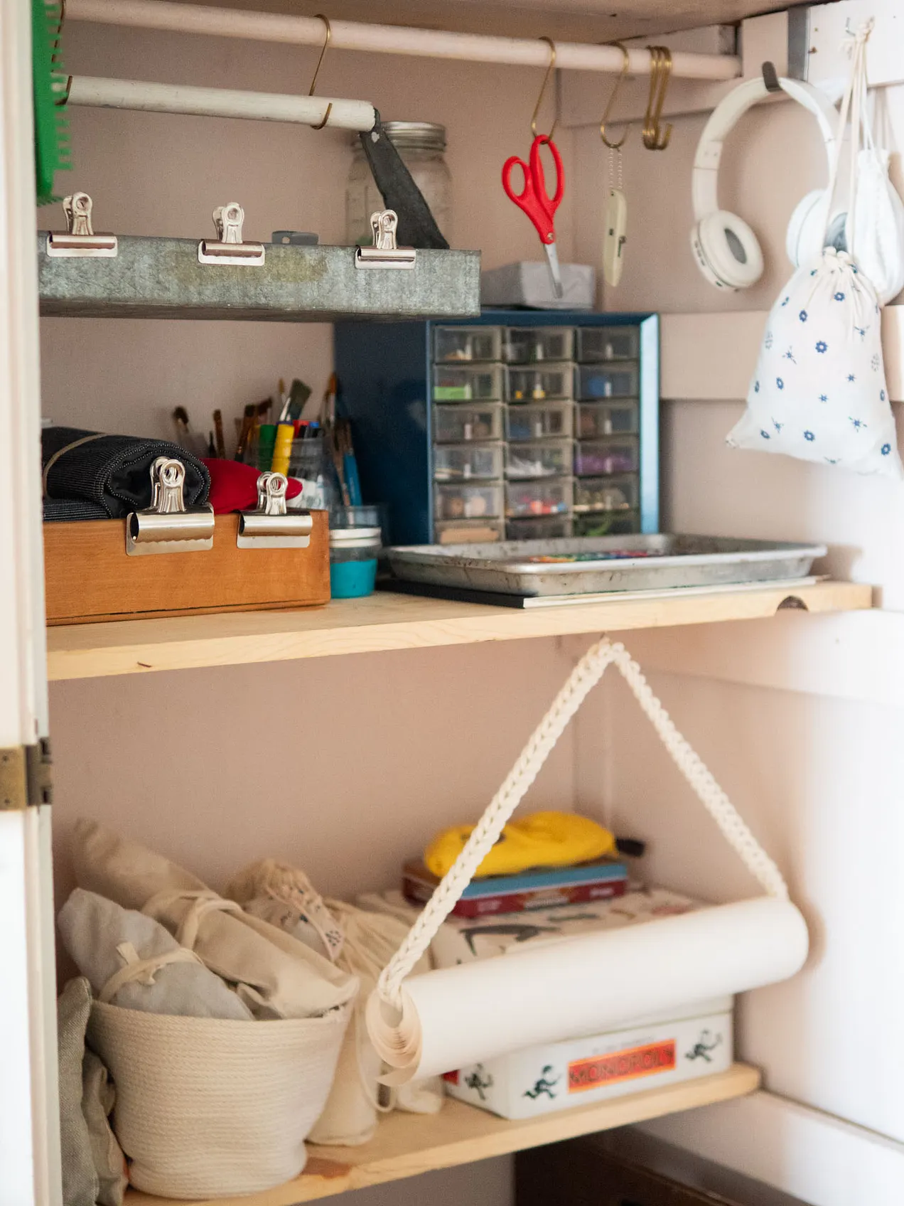 Interior of an open storage cabinet or closet organized with art supplies. Upper shelves hold binder clips, paint brushes, scissors, a small multi-drawer organizer, and metal trays. A white rope basket and a rolled paper carrier sit on the lower shelf alongside fabric pouches and small notebooks. White headphones and a small drawstring bag hang from hooks on the right side.