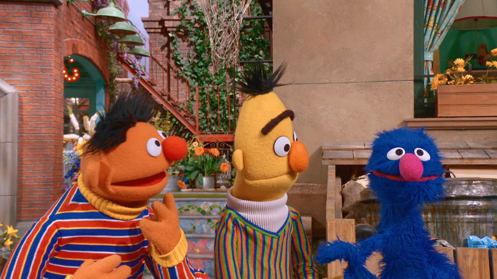 A screenshot from an episode of Sesame Street showing Ernie (in his classic orange-and-yellow horizontal striped shirt), Bert (in a vertical multicolor striped shirt), and Grover (in blue fur) standing together on the Sesame Street set.