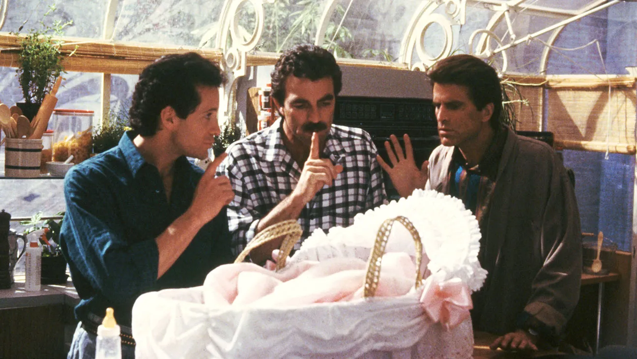 A scene from the 1987 film Three Men and a Baby showing three men — one in a blue shirt, one in a plaid shirt with a mustache, and one in a gray robe — huddled together with concerned expressions around a baby in a white wicker bassinet decorated with pink ruffles and a bow.