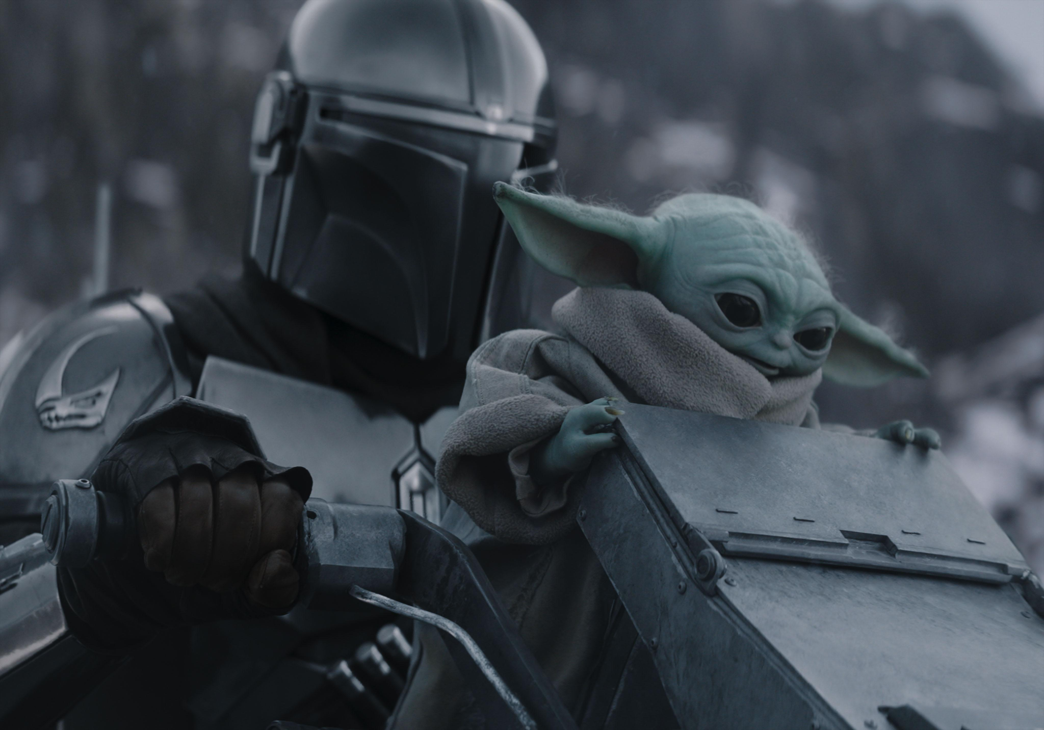 A close-up scene from The Mandalorian showing a helmeted armored figure in silver Mandalorian armor alongside Grogu (commonly known as Baby Yoda), a small green creature with large ears and dark eyes, peering over the edge of a metal surface. The image has a cool, desaturated blue-gray tone.