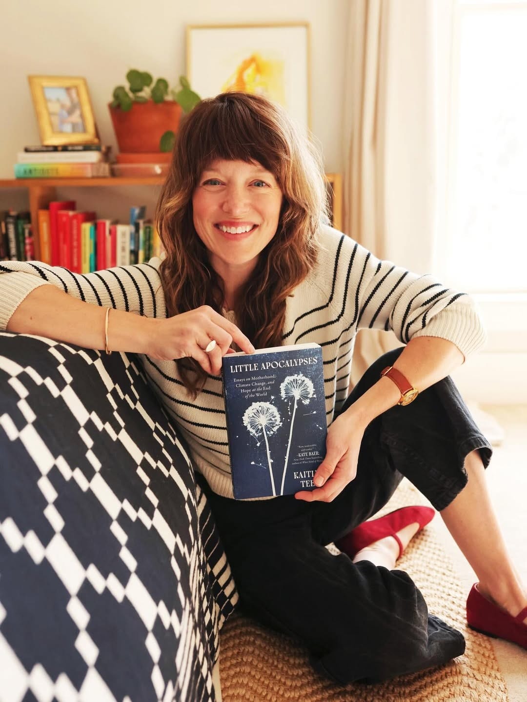 A smiling woman with wavy brown hair and bangs sits on the floor, leaning on a bed with a blue and white geometric patterned bedspread. She is holding up a copy of Little Apocalypses: Essays on Motherhood, Climate Change, and Hope at the End of the World. She wears a black-and-white striped sweater, black pants, and red shoes. Bookshelves and a potted plant are visible behind her.