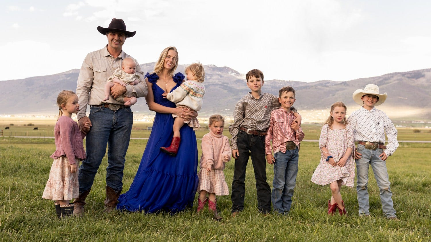 A father in a cowboy hat holding an infant and a mother in a blue dress holding a toddler in red boots, surrounded by seven children in coordinated pastel and western wear, standing in a grassy field with mountains in the background.