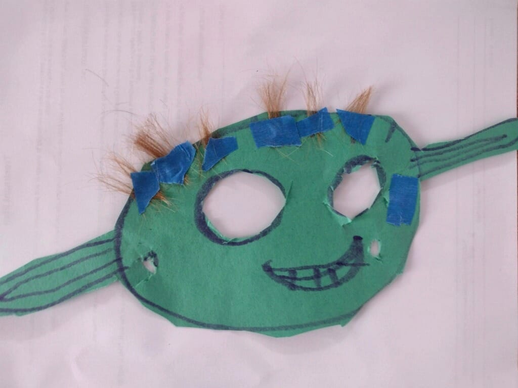 A child's handmade Yoda mask crafted from green construction paper, with two cut-out eye holes and a marker-drawn smiling mouth with teeth. Tufts of real hair are attached above the eyes with strips of blue tape, and long pointed paper ears with drawn-on lines extend from each side of the head.