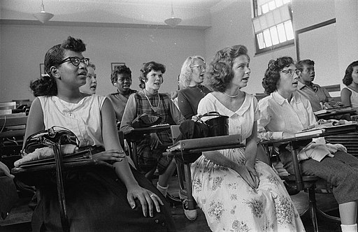 Black and white photograph of several female students, both white students and Black, wearing 1950s era clothing and seated at individual desks. They all look in the same direction toward a point outside the frame. Their mouths are open as in song or recitation.