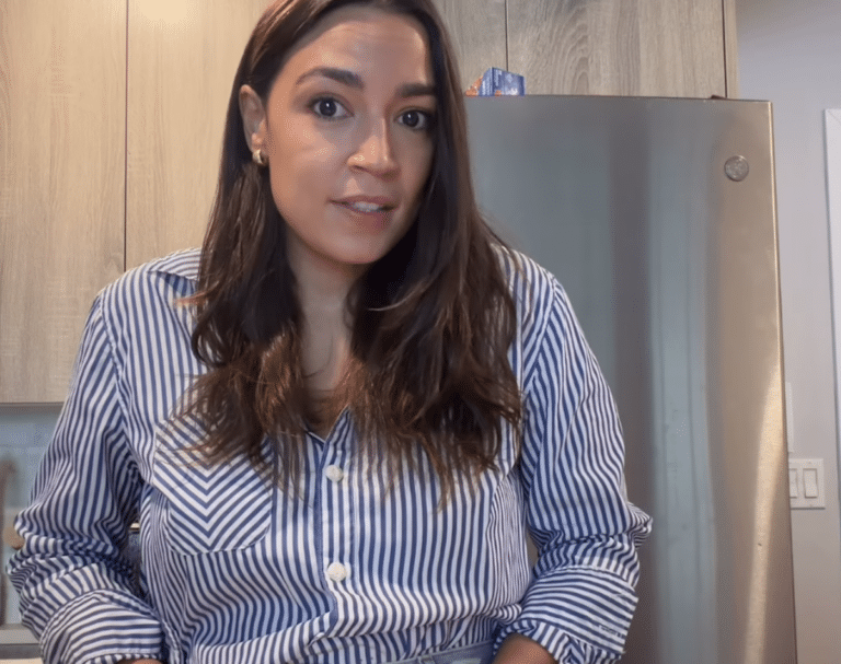 Alexandria Ocasio-Cortez speaking to camera during a video about freezing her eggs, wearing a blue and white striped button-down shirt, kitchen visible in background.