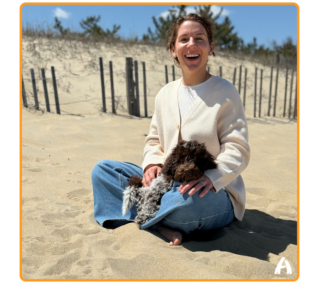A woman sits cross-legged on a sandy beach, laughing and cradling a small curly-haired dog in her lap.