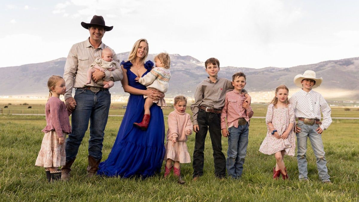 A father in a cowboy hat holding an infant and a mother in a blue dress holding a toddler in red boots, surrounded by seven children.