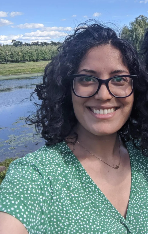 Sri Juneja, a woman with dark curly hairbehind her is a calm pond bordered by grass and a tree line, under a blue sky with scattered white clouds.