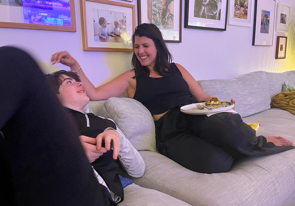 A smiling woman sits on a gray couch with a plate of food, playfully touching the hair of a teenager who is reclining next to her.