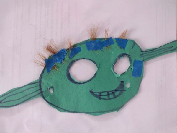 A child's handmade Yoda mask crafted from green construction paper, with two cut-out eye holes and a marker-drawn smiling mouth with teeth. Tufts of real hair are attached above the eyes with