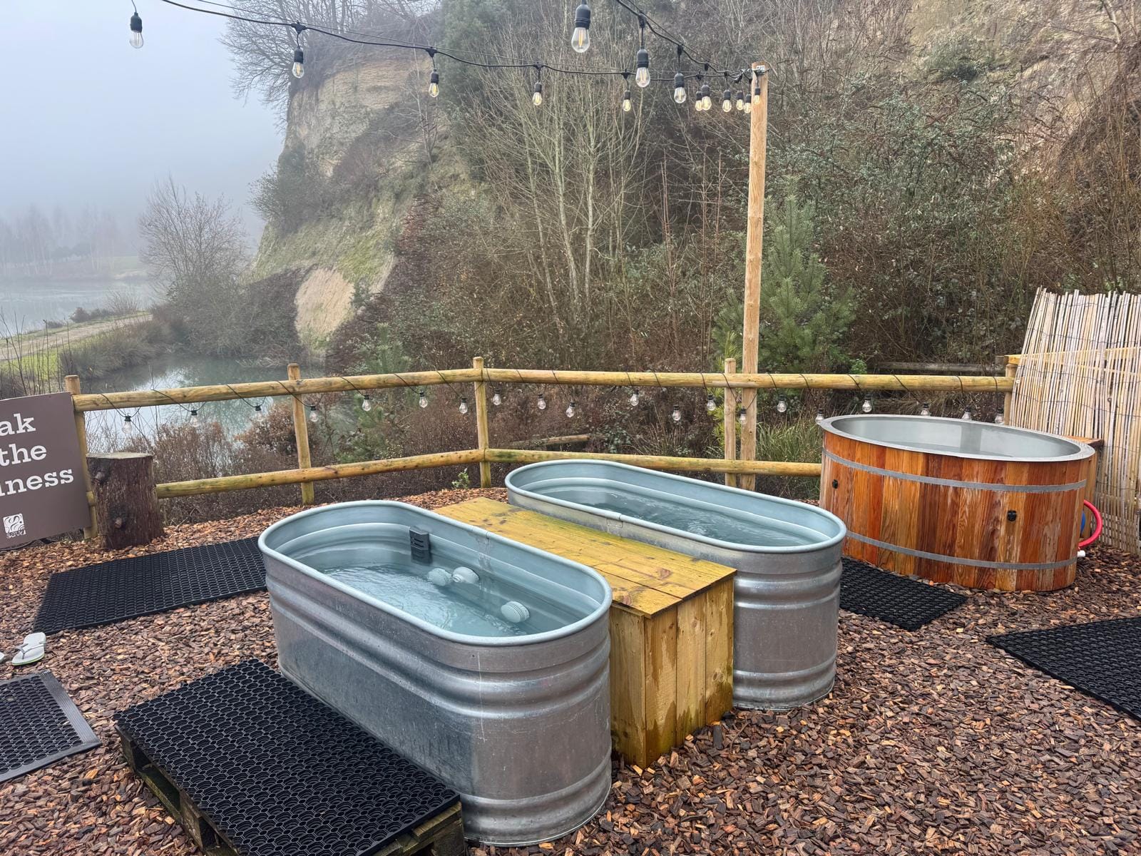 Three cold plunge tubs at Savu sauna overlooking Buckland Park Lake, with rubber mats on bark ground.