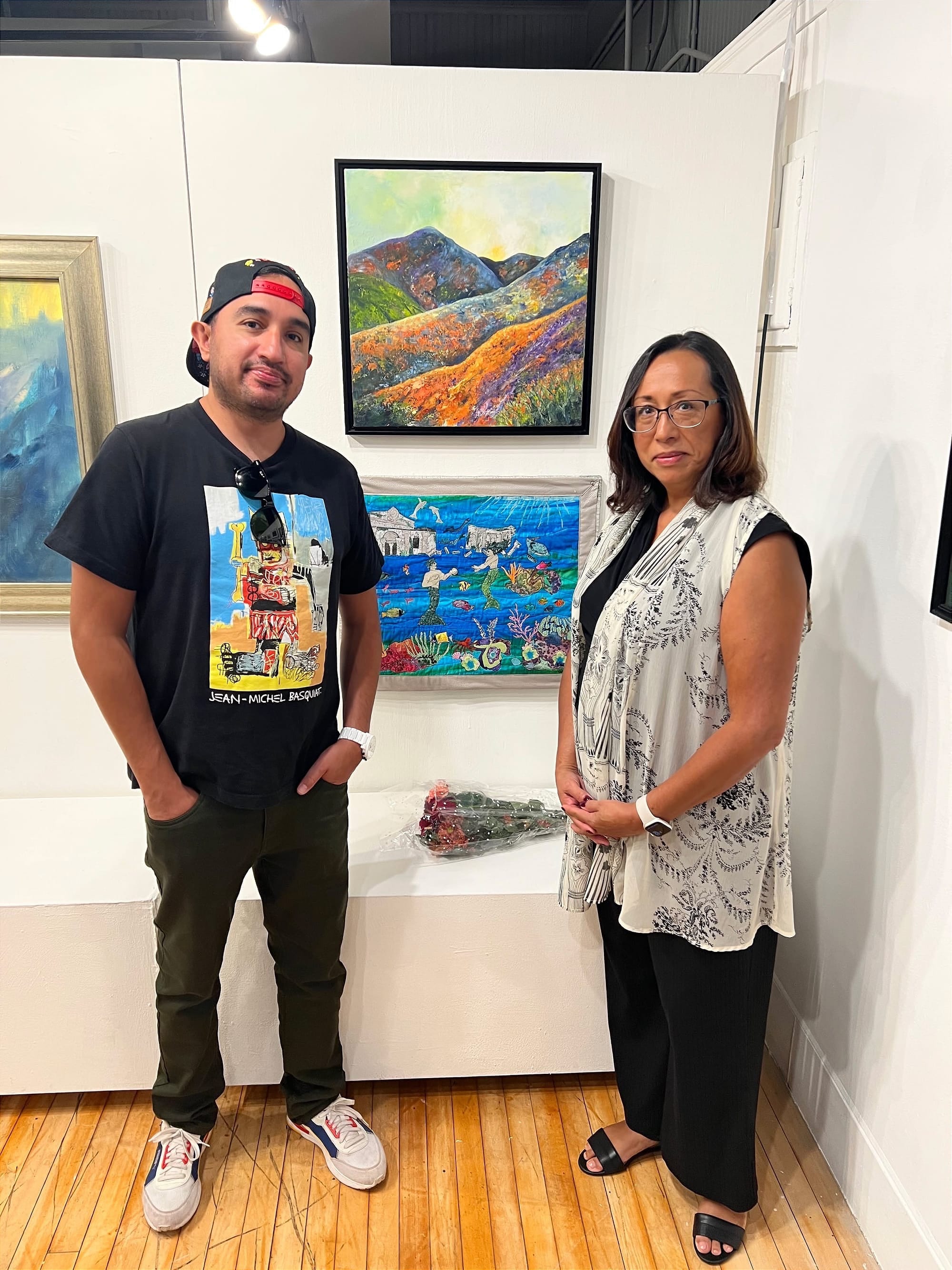 A young man and his mother stand side by side, a bouquet of roses between them, and pictures hanging on a gallery wall behind them.