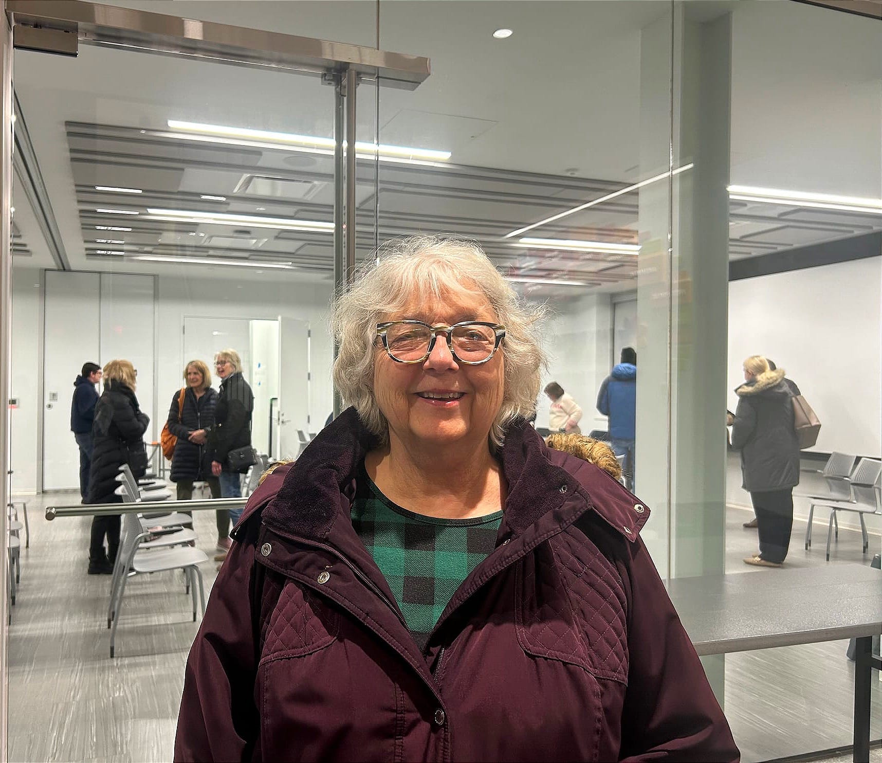 A woman with gray hair and glasses wearing a maroon quilted jacket smiles at the camera inside a community meeting space, with other attendees visible in the background.