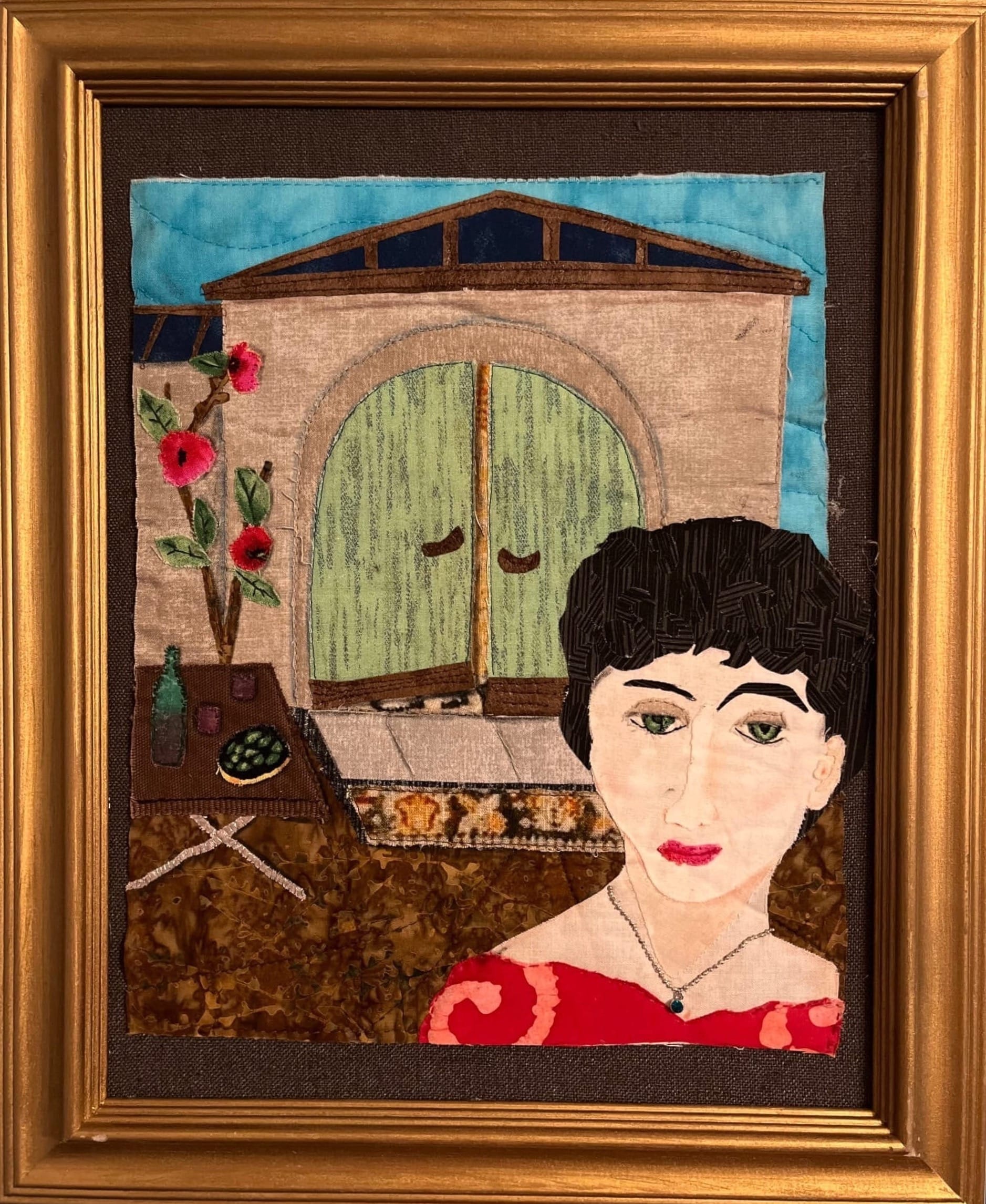 Fabric appliqué artwork titled 'GiGi' by Piotrowski, depicting a dark-haired woman in a red dress in front of a building with an arched green door, flanked by pink flowers and a small table with a bottle and bowl.