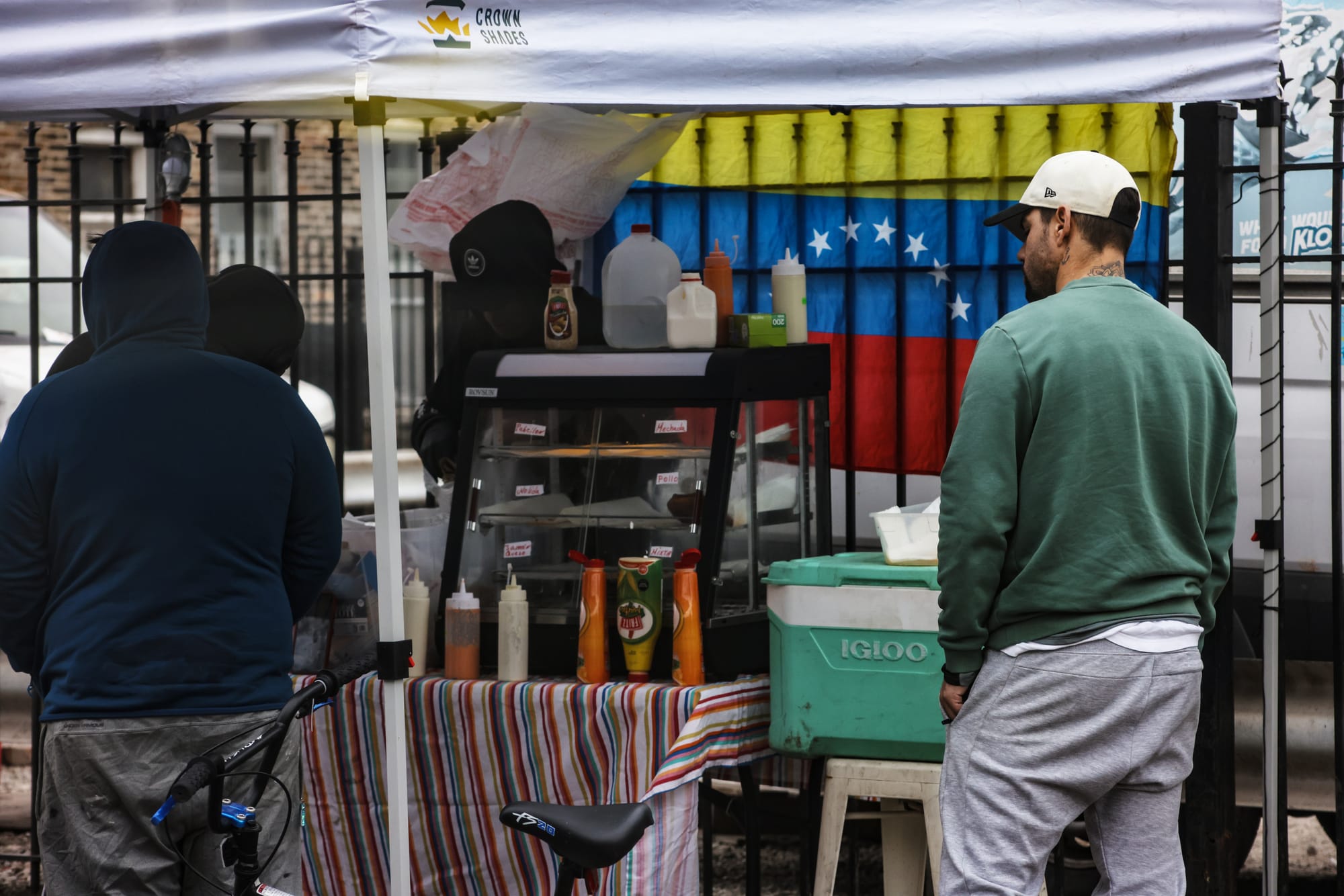 Three people wait at an outdoor food vendor stand beneath a white tent, with a Venezuelan flag displayed behind the food display case.