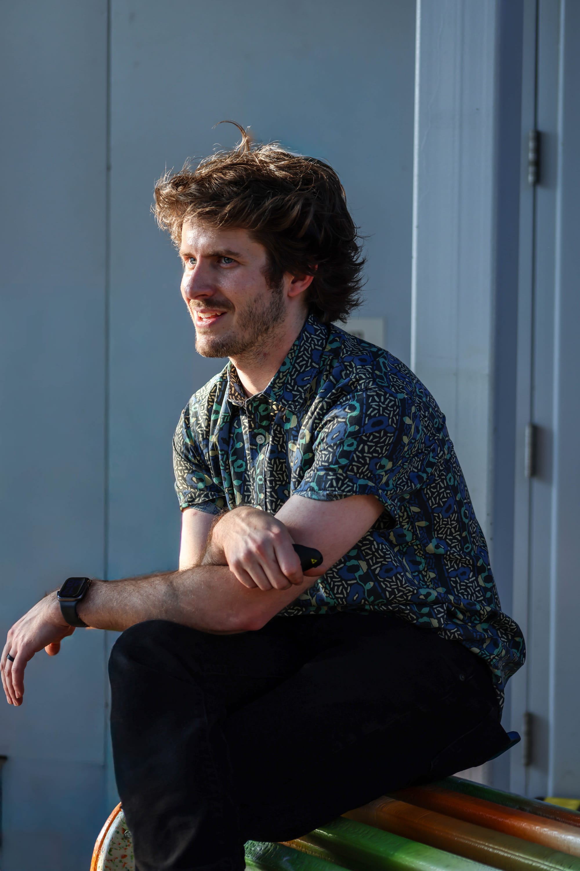 Man [Cody Norman] in a vibrant floral shirt, seated, legs crossed against a grey background.