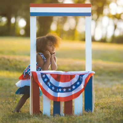 A child in red white and blue dress leaning against a red white and blue pallet stand with a flag inspired banner.