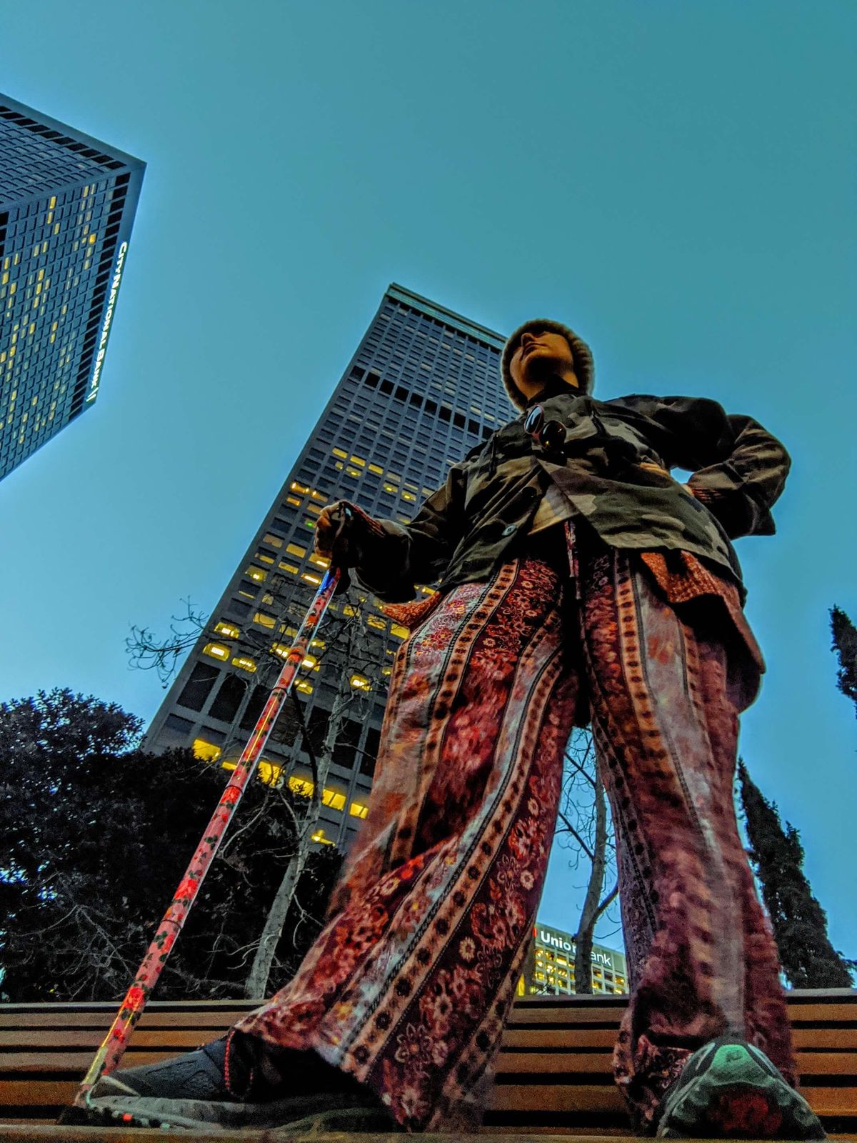 shot of PJ standing against skyscrapers, wearing sneakers and patterened pants with her pink floral cane. she's wearing a army fatigue jacket and a hat.