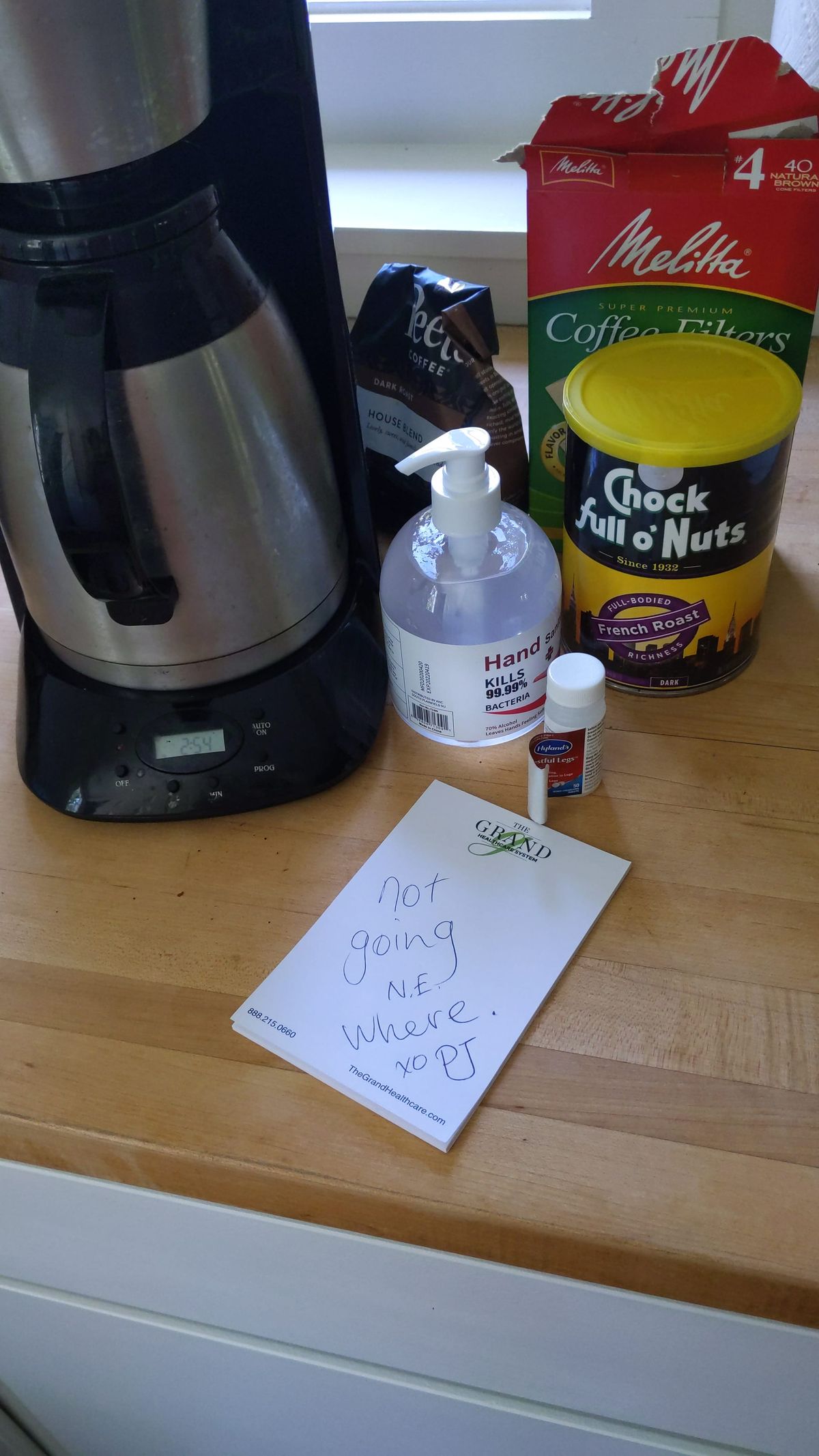 on a wood counter: a black and titanium coffee pot, a bottle of hand sanitizer, coffee filers, a chock full of nuts can, a white pad with handwriting "not going N.E. Where xo PJ"
